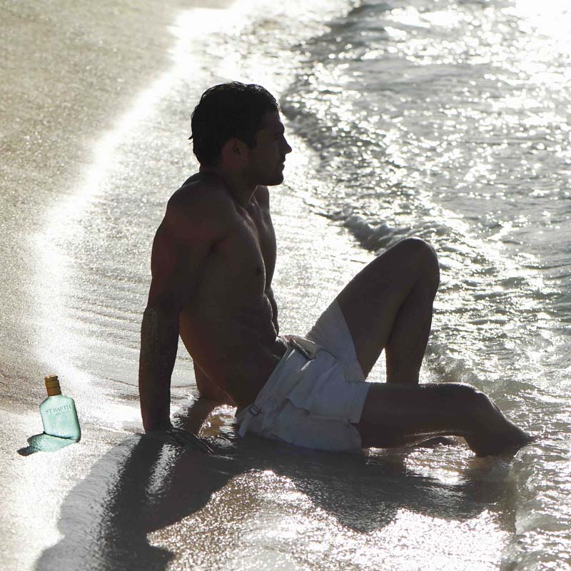 Lifestyle shot of Edit website SEO Ligne St. Barth Blue Lagoon Shower Gel 6.8 oz next to man sitting on the beach shoreline with wave coming in