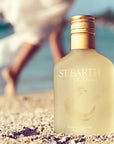 Lifestyle shot of Ligne St. Barth Premium Care Oil 6.8 oz in the sand with woman walking along the beach in the background