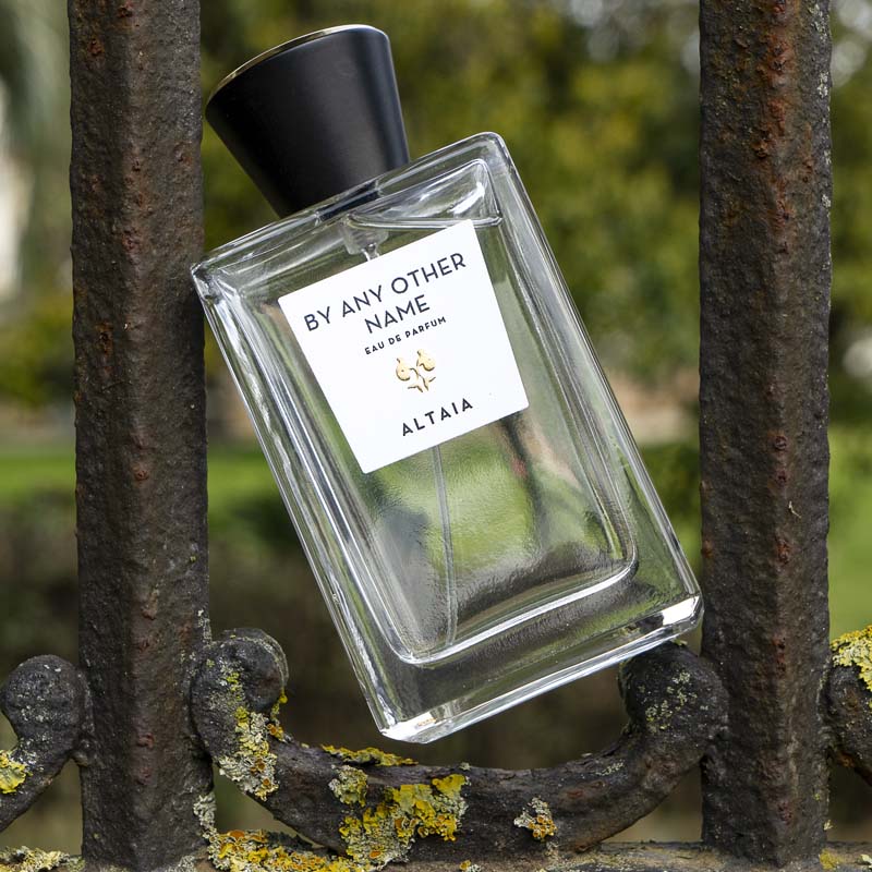 Beauty shot of ALTAIA By Any Other Name Eau de Parfum placed in between bars of an old metal gate with rust and moss