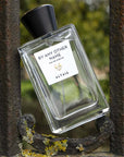 Beauty shot of ALTAIA By Any Other Name Eau de Parfum placed in between bars of an old metal gate with rust and moss