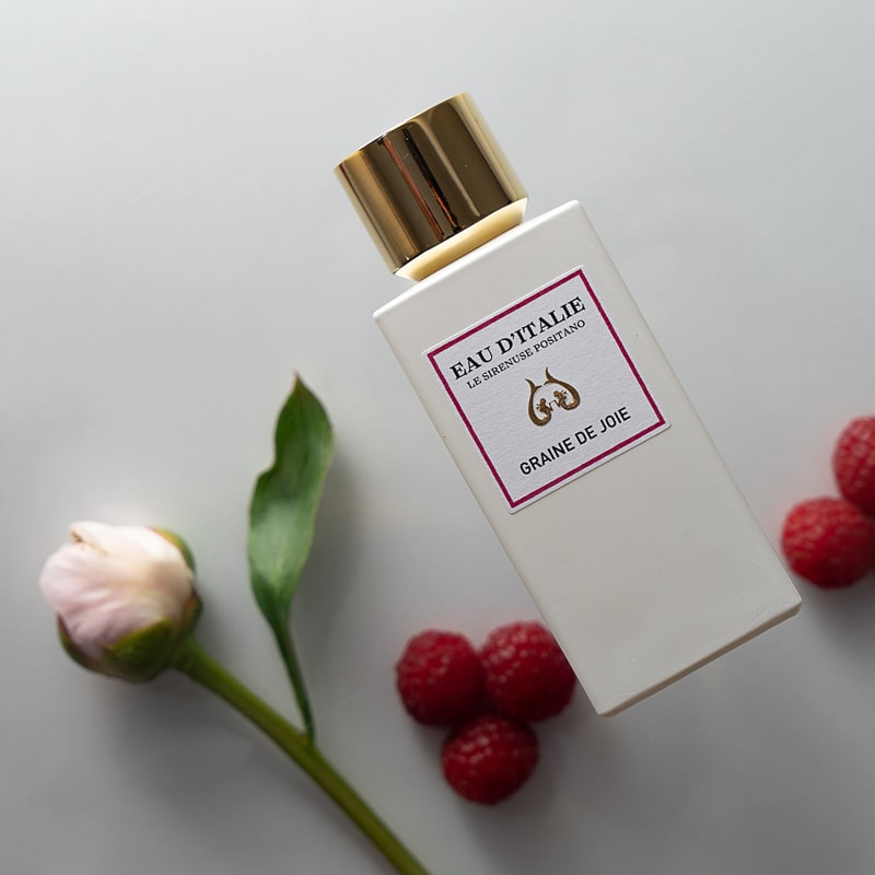 Lifestyle shot top view of Eau d&#39;Italie Graine de Joie Eau de Parfum Spray (100 ml) with raspberries and single pink flower in the background