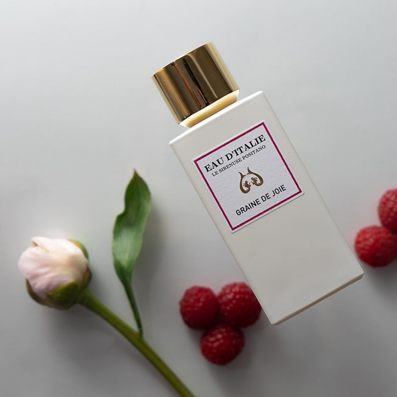 Lifestyle shot top view of Eau d'Italie Graine de Joie Eau de Parfum Spray (100 ml) with raspberries and single pink flower in the background