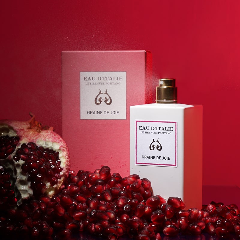 Lifestyle shot of Eau d&#39;Italie Graine de Joie Eau de Parfum Spray (100 ml) with box and pomegranate and seeds in the foreground