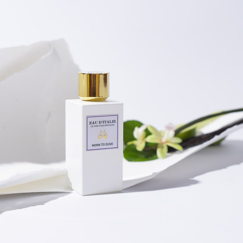 Lifestyle shot of Morn to Dusk Eau de Parfum Spray (100 ml) with white torn paper and white flowers in the background