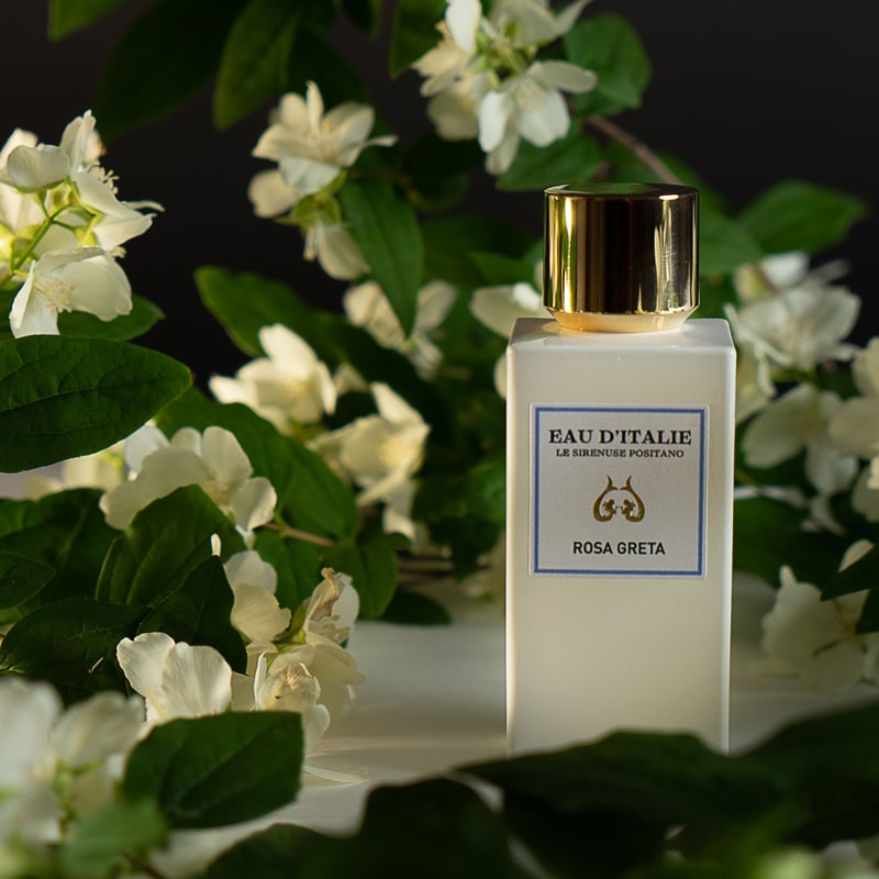 Lifestyle shot of Eau d&#39;Italie Rosa Greta Eau de Parfum Spray bottle (100 ml) with white flowers in the background