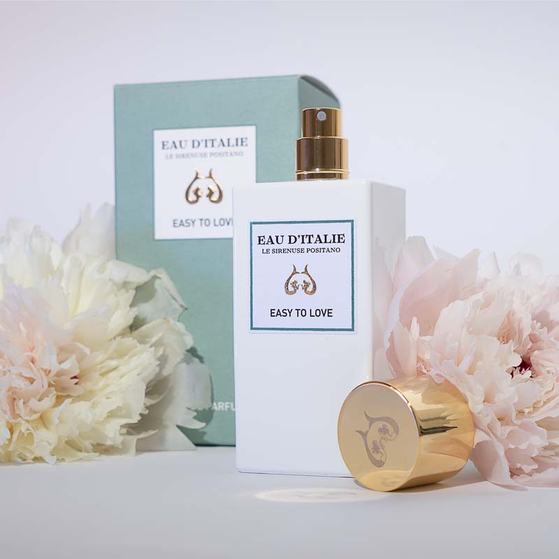Lifestyle shot of Eau d'Italie Easy to Love Eau de Parfum Spray (100 ml) with box cap off and pale pink and cream flowers in the background and foreground