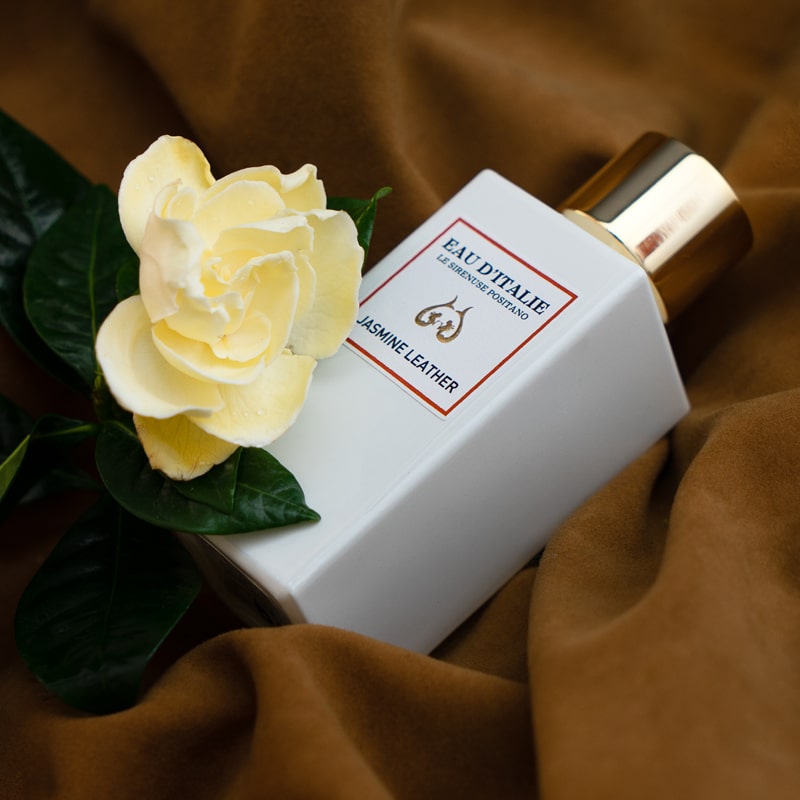 Lifestyle shot of Eau d&#39;Italie Jasmine Leather Eau de Parfum Spray (100 ml) on brown fabric with jasmine flower