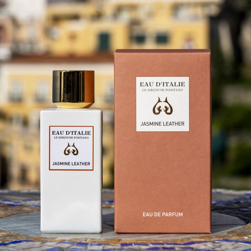 Lifestyle shot of Eau d&#39;Italie Jasmine Leather Eau de Parfum Spray (100 ml) with box on tiled mosaic table and the Positano hillside in the background